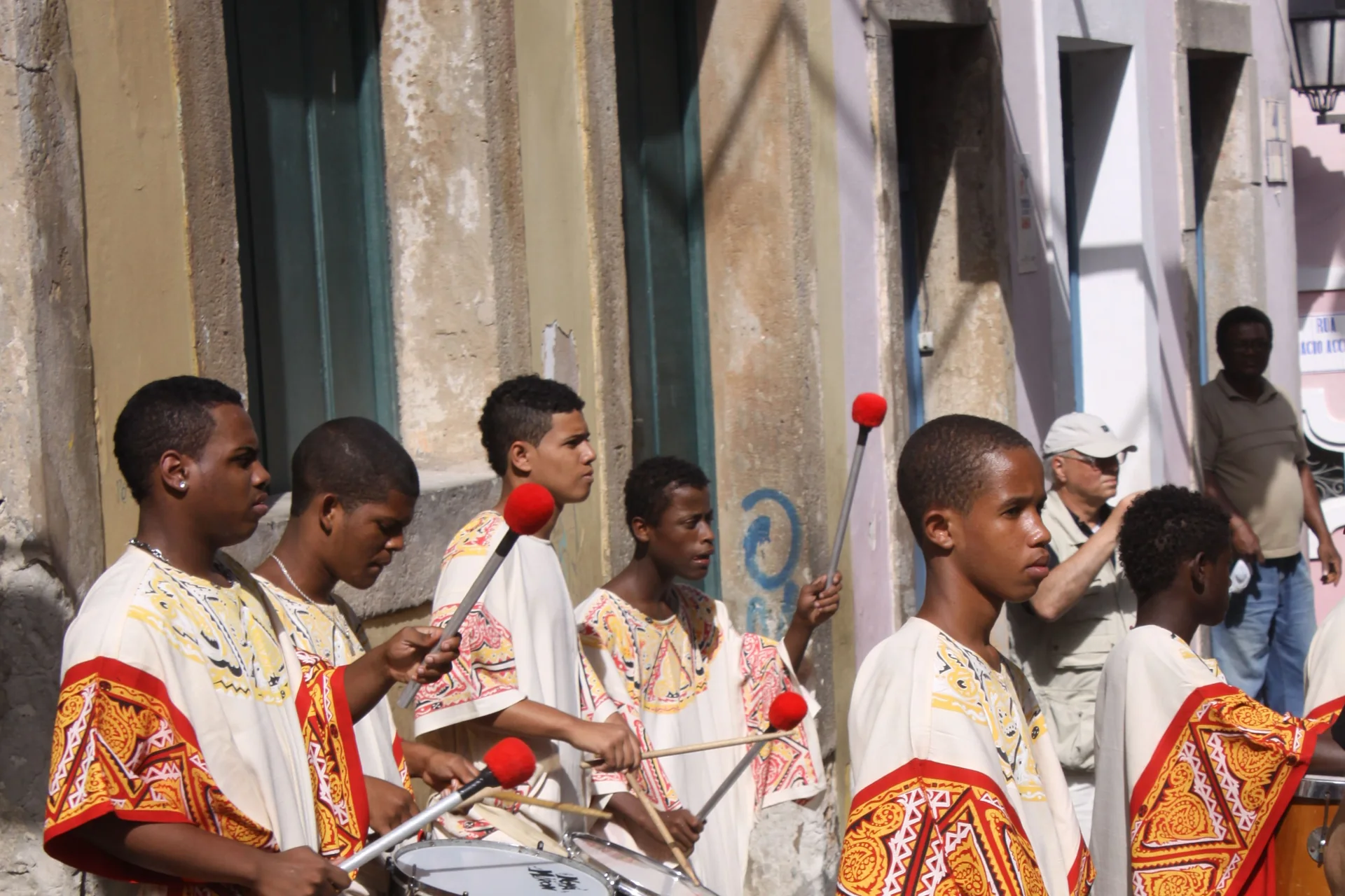 A drum corps in yellow, white and red costume plays large parade drums on a cobbled street lined with colourful colonial buildings in the Pelourinho, Salvador, Brazil