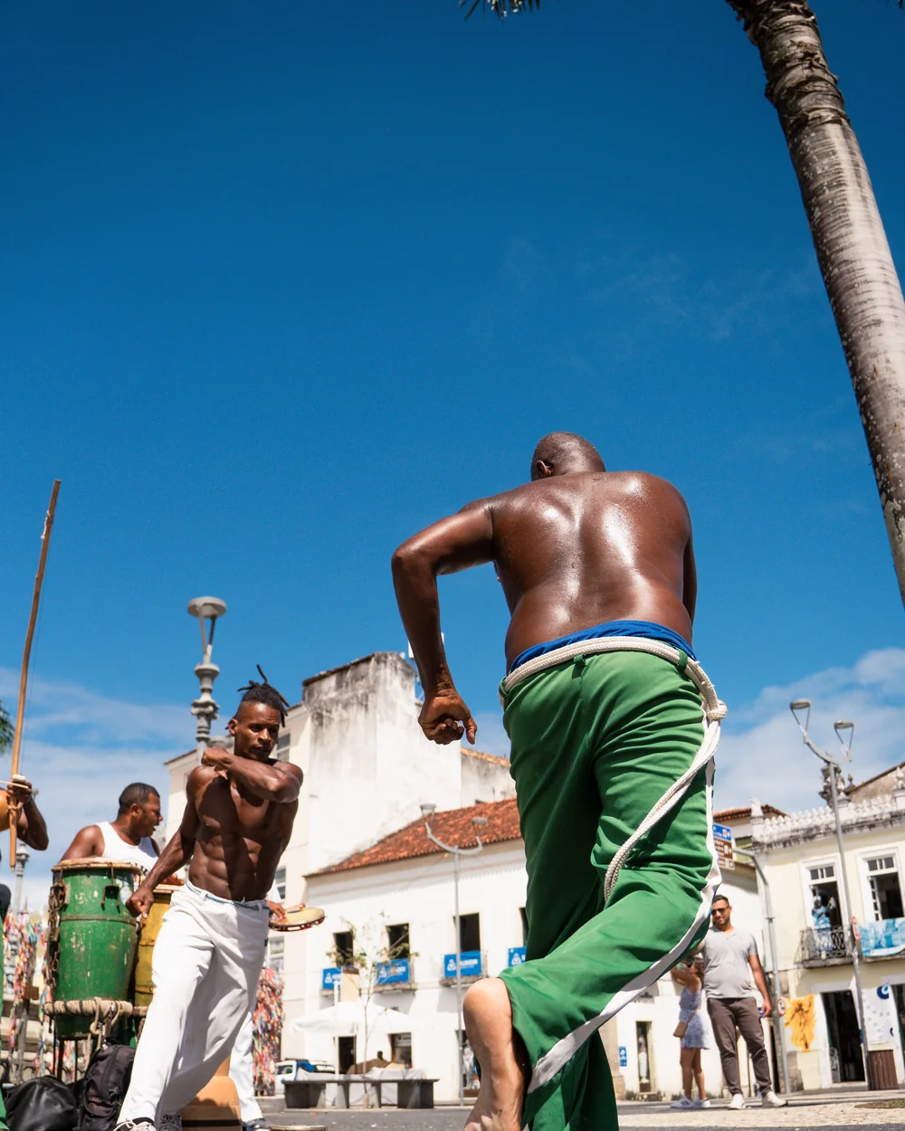 A capoeirista mid-cartwheel in a Pelourinho-style square, drummers and colonial buildings behind (representative photograph of the folklore company’s energy, not a maculelê stick routine)
